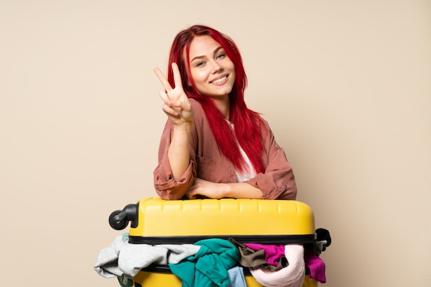 Mujer del viajero con una maleta llena de ropa aislado en la pared de color beige sonriendo y mostrando el signo de la victoria
