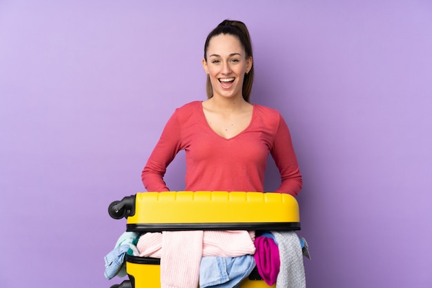 Mujer del viajero con una maleta llena de ropa sobre la pared púrpura aislada con expresión facial sorpresa