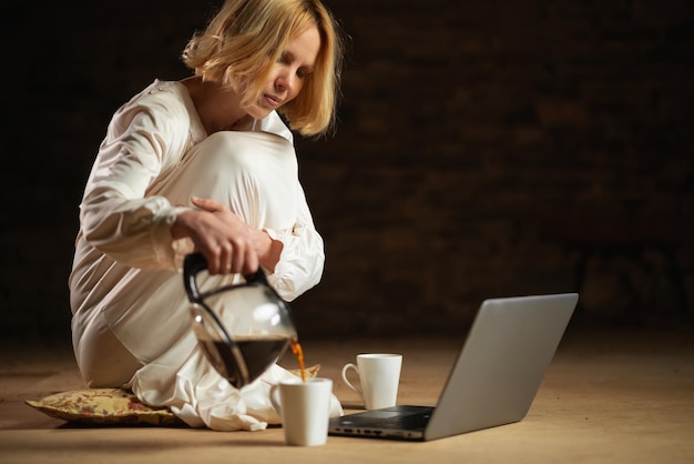 Una mujer vierte café mientras está sentada frente a una computadora portátil. Habitación vacía. Fondo oscuro. Fecha límite para reciclar el trabajo duro.