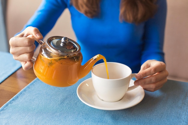 Una mujer vierte té de hierbas vitamínicas y saludables en una taza blanca vacía en un café en un frío día de otoño