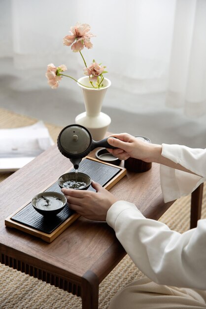 Una mujer vierte té en una taza sobre una mesa frente a un jarrón con flores.