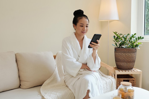 Mujer vietnamita joven positiva en bata de baño descansando en el sofá después del procedimiento de spa y leyendo noticias en