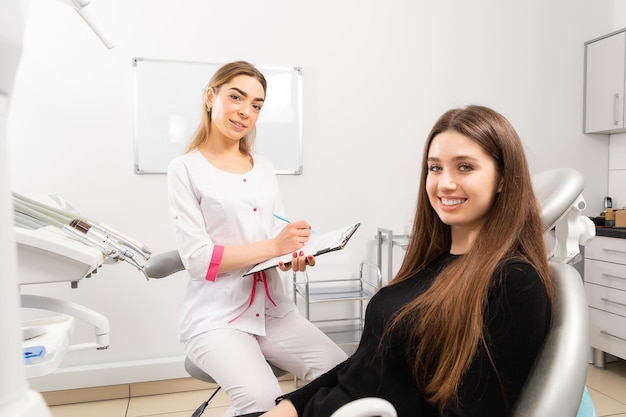 Una mujer vino al dentista. El médico llena un formulario y realiza una encuesta a los pacientes. Todo el mundo está sonriendo. Están ubicados en una oficina de dentista blanca y moderna.