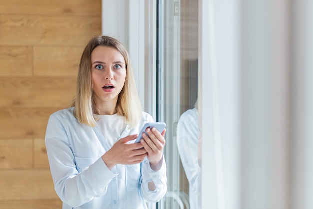 Mujer vio malas noticias en el teléfono inteligente de pie junto a la ventana en la oficina o en casa