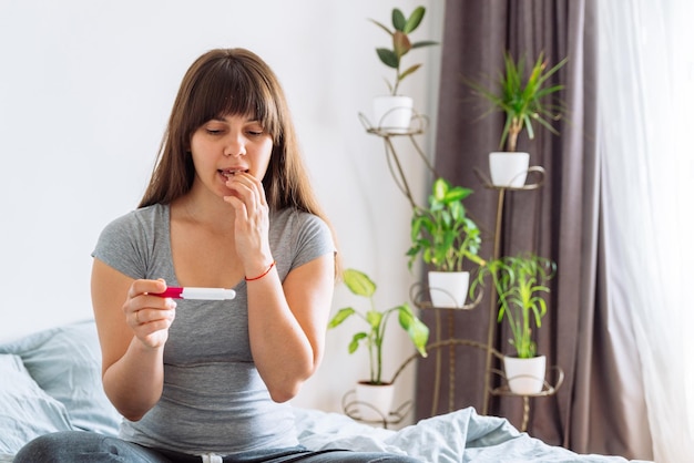 Mujer con vista preocupada mirando la prueba de embarazo mientras está sentada en la cama