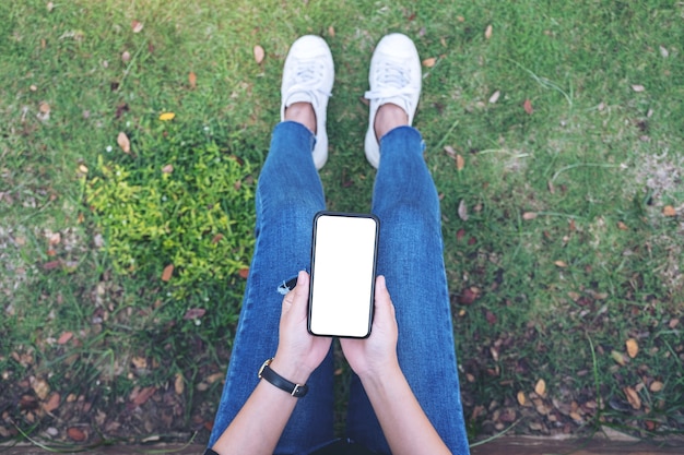 Mujer de la vista superior que sostiene el teléfono móvil negro con la pantalla en blanco en blanco mientras está sentado en el parque