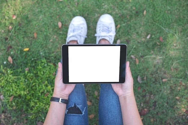 Mujer de vista superior sosteniendo y usando tablet pc negro con pantalla blanca en blanco horizontalmente mientras está sentado en el parque