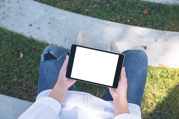 Mujer de vista superior sosteniendo y usando tablet pc negro con pantalla de escritorio blanca en blanco horizontalmente mientras está sentado en el parque