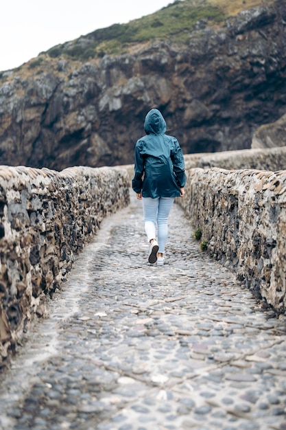 Mujer vistiendo un impermeable caminando por un camino pavimentado rodeado por dos muros de piedra