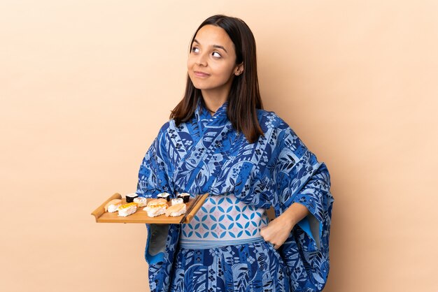 Mujer vistiendo kimono y sosteniendo sushi aislado haciendo dudas gesto mirando hacia el lado
