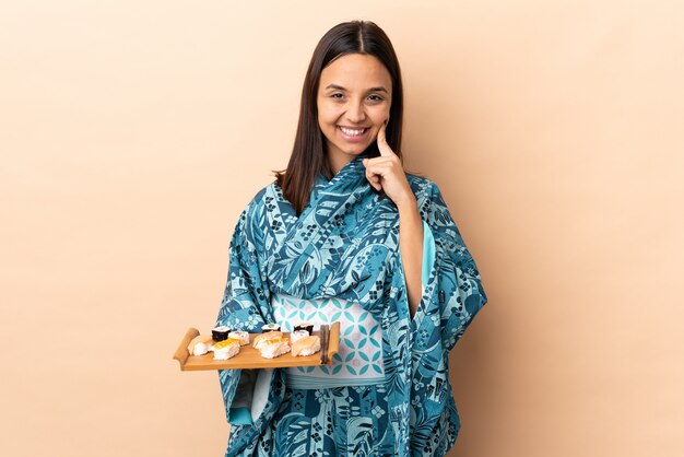 Mujer vistiendo kimono y sosteniendo sushi sobre la pared sonriendo con una expresión feliz y agradable