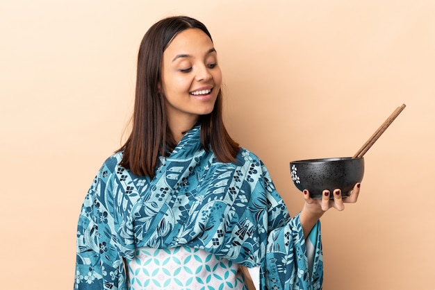 Mujer vistiendo kimono y sosteniendo un tazón lleno de fideos con expresión feliz mientras sostiene un tazón de fideos con palillos