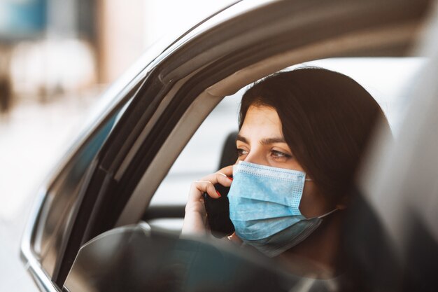 Mujer vistiendo una máscara médica estéril en un taxi en un asiento trasero mirando por la ventana hablando por teléfono