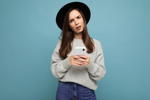 Mujer vistiendo sombrero negro y suéter gris sosteniendo smartphone mirando a la cámara aislada sobre fondo