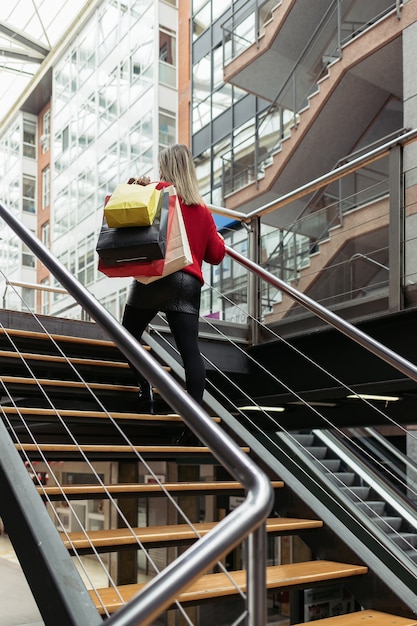 Mujer vistiendo un suéter rojo subiendo las escaleras de un centro comercial llevando bolsas de compras.