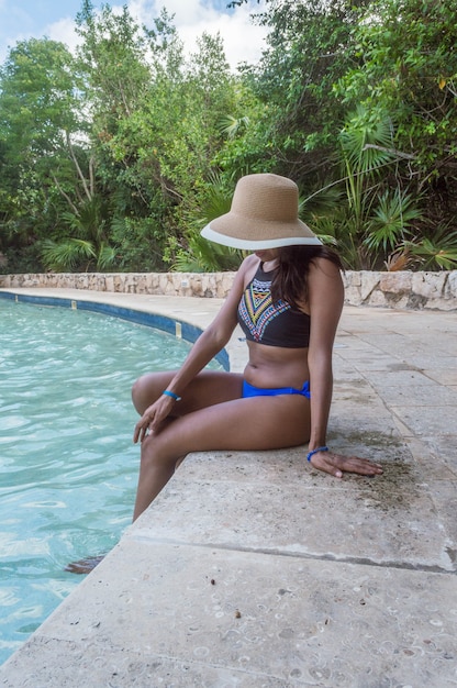 Mujer vistiendo traje de baño con sombrero marrón sentado en el borde de la piscina mientras mira el agua