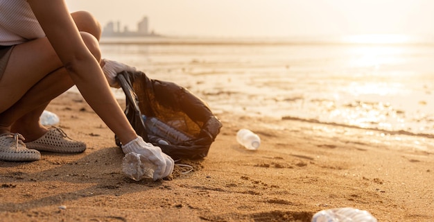 Mujer voluntaria recogiendo una botella de plástico en una bolsa de basura negra para limpiar la playa