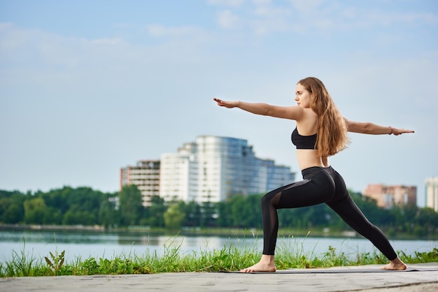 Mujer de yoga parada en una pose guerrera Virabhadrasana cerca del río en el fondo de edificios urbanos con espacio para copiar