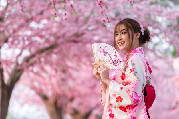 Mujer en yukata (vestido de kimono) sosteniendo abanico plegable y mirando flor de sakura o flor de cerezo que florece en el jardín