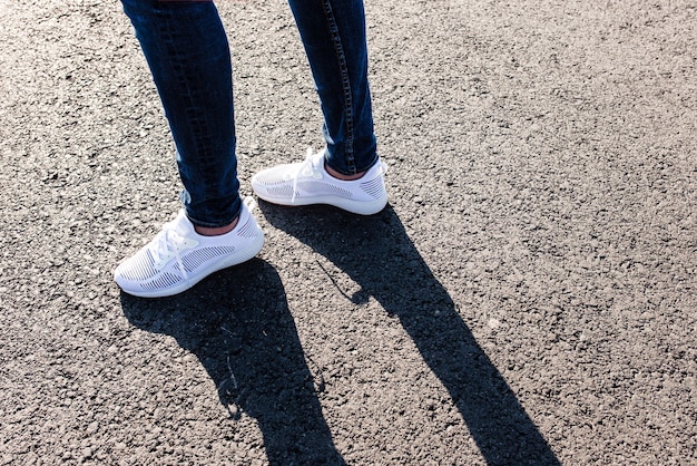 Mujer en zapatillas blancas de pie sobre la carretera asfaltada hacia el sol, de cerca.