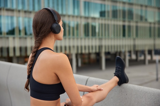 Mujer en zapatillas de deporte negras recortadas estira las piernas al aire libre en un ambiente moderno se calienta antes de hacer ejercicio plantea en el puente cerca del edificio de cristal disfruta de la banda sonora favorita