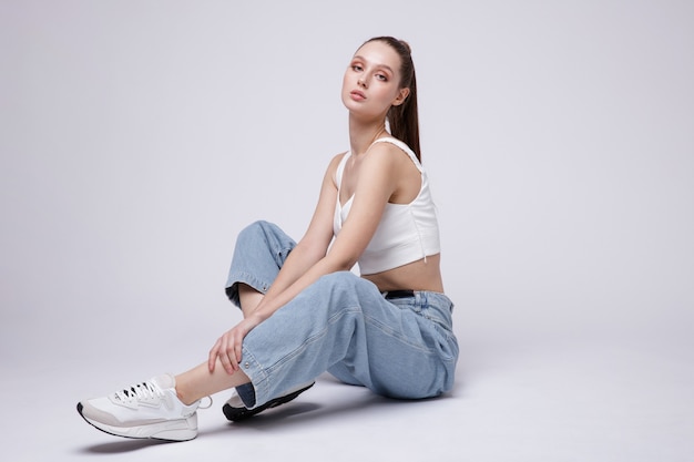 Mujer en zapatillas de deporte superior blanco jeans de mezclilla azul sobre fondo blanco Studio Shot modelo está sentado