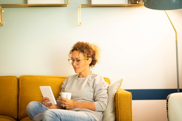 Las mujeres adultas tienen actividades de ocio relajantes en casa sentadas en el sofá y leyendo un libro electrónico con una tableta lectora electrónica Las mujeres usan la conexión de tecnología de Internet en el sofá