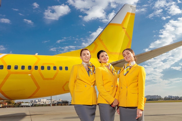 Mujeres alegres azafatas en uniforme de aerolínea mirando a la cámara y sonriendo mientras están de pie cerca del avión contra el cielo nublado