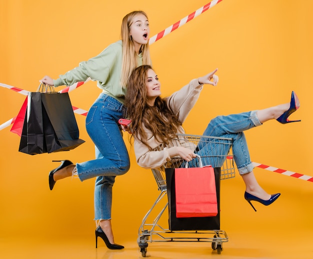 Mujeres apuntando a copyspace con carrito lleno de bolsas de compras y cinta de señal aislada sobre amarillo