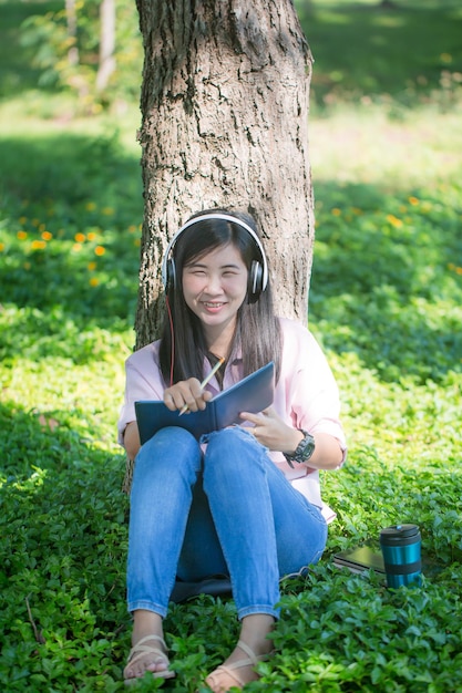 Las mujeres asiáticas escuchan música con auriculares y trabajan con computadoras portátiles debajo de un árbol en un día soleado