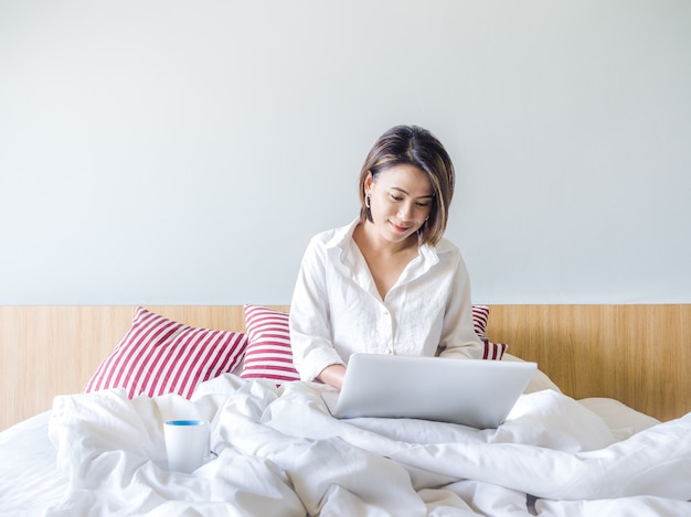 Mujeres asiáticas hermosas con el pelo corto que lleva la camisa blanca que trabaja con la computadora portátil en la cama en la casa.