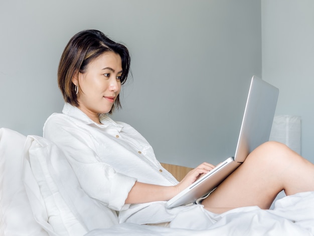 Mujeres asiáticas hermosas con el pelo corto que lleva la camisa blanca que trabaja con la computadora portátil en la cama en la casa.
