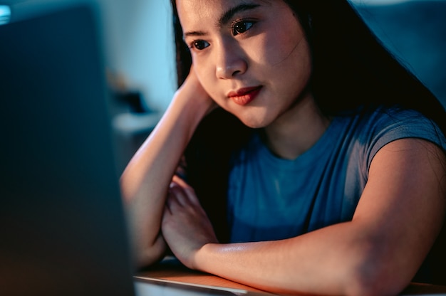 Mujeres asiáticas jóvenes que trabajan con la computadora portátil en la sala de estar en casa en la oscuridad en la lámpara