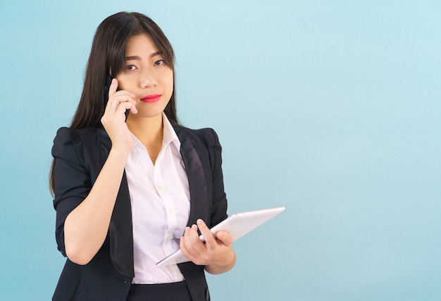 Las mujeres asiáticas jóvenes en traje de pie usando su teléfono y sosteniendo una tableta digital contra el fondo azul.