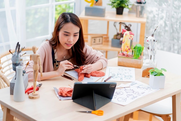 Las mujeres asiáticas pasan el fin de semana para su curso en línea de escultura de arcilla de hobby en casa, adultos jóvenes que estudian desde un curso de transmisión de tabletas en línea en un estilo de vida informal asiático disfrazado de delantal en casa