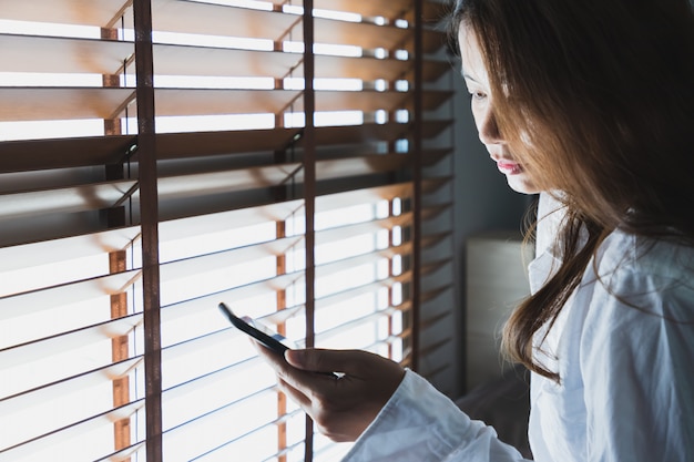 Las mujeres asiáticas que usan una camisa blanca están mirando el teléfono en la ventana en el dormitorio