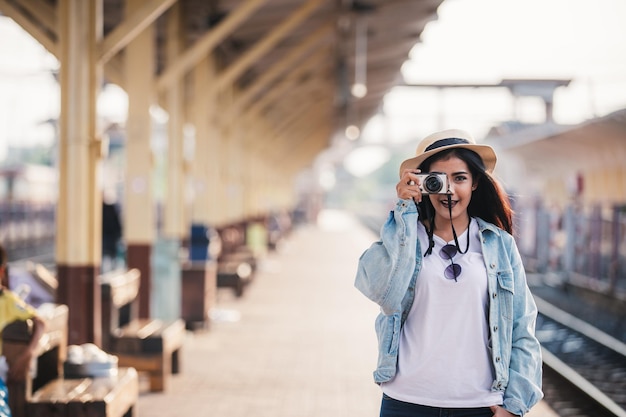 Las mujeres asiáticas sonríen con una cámara de viaje de vacaciones en la estación de tren con un viajero relajante Conc