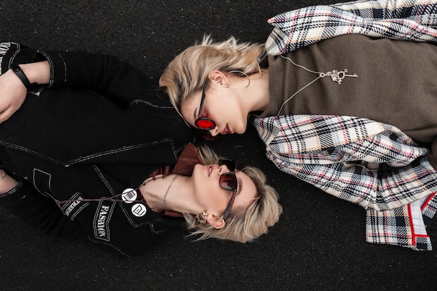 Mujeres atractivas de moda en ropa con estilo vintage en gafas de sol de moda se relajan en el asfalto y se miran