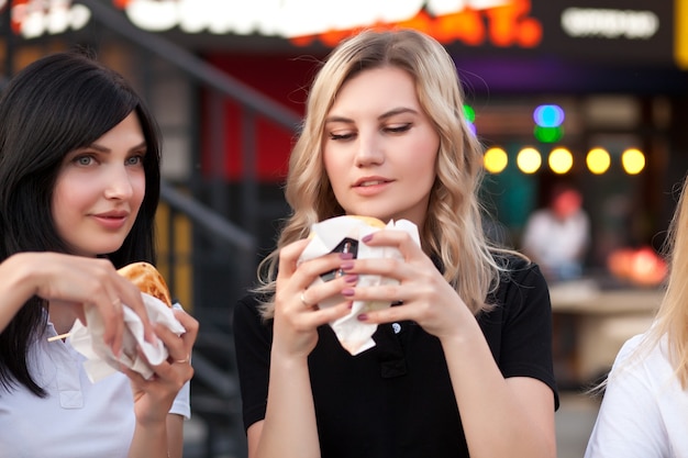 Mujeres bastante jóvenes comiendo hamburguesas al aire libre en la calle.