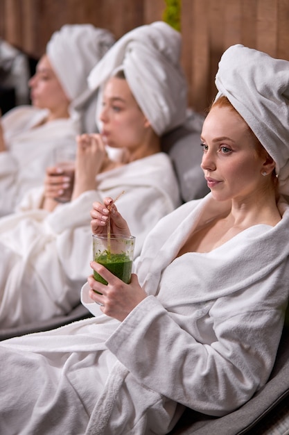 Mujeres en batas de baño que pasan tiempo en el salón del hotel del balneario, disfrutando de jugos orgánicos naturales, después de los procedimientos de belleza. Concepto de cuidado, bienestar y relajación corporal. centrarse en la mujer pelirroja con una toalla en la cabeza