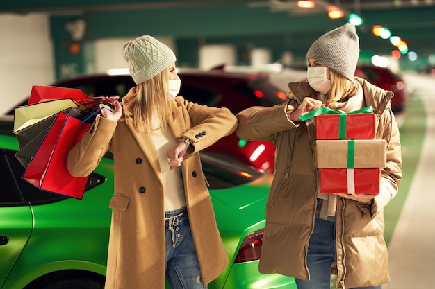 Mujeres con bolsas de la compra y regalos de Navidad en máscaras en el estacionamiento subterráneo