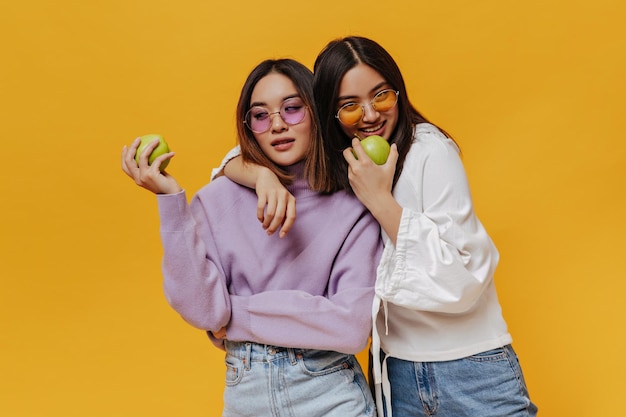 Mujeres bonitas asiáticas con gafas de sol coloridas sostienen manzanas verdes frescas Chicas jóvenes con sudaderas posan sobre fondo naranja