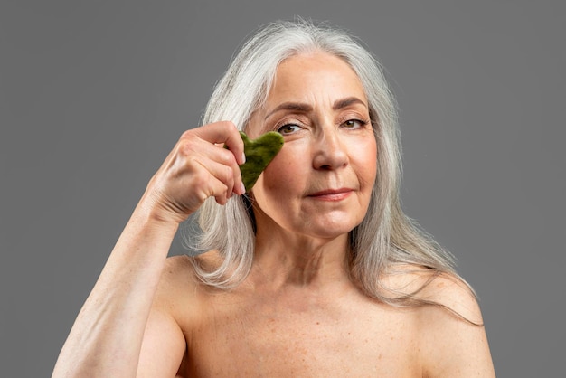 Las mujeres caucásicas maduras con cabello gris hacen masajes con piedras de jade en casa aisladas en un estudio de fondo gris