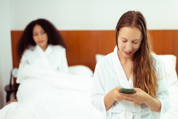 Las mujeres caucásicas y negras se aman juntas y se acuestan en la cama como personas de diversidad Adulto joven Pareja romántica revisa el teléfono inteligente detrás de su espalda en busca de texto de chat secreto como espacio de copia de sentimiento celoso