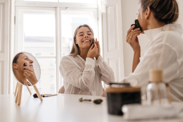 Mujeres caucásicas positivas haciendo masaje facial con masajeador de jade alisando las arrugas en el interior. Concepto de tratamientos de belleza y spa para el cuidado de la piel.