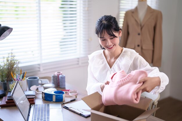 Mujeres comerciantes asiáticas jóvenes comprobando compras en línea en la computadora portátil y empaquetando productos de ropa en paquetes para el envío de entrega para el cliente mientras trabajan en negocios de compras en línea de la oficina en casa