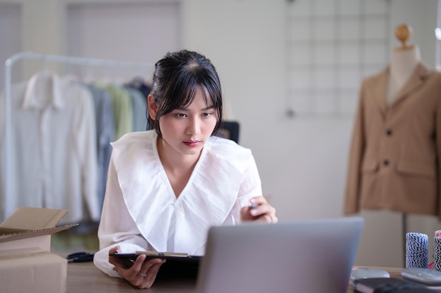 Foto mujeres comerciantes asiáticas jóvenes comprobando compras en línea, pedidos y información del cliente en la computadora portátil para tomar notas en el portapapeles mientras trabajan y envían entregas para negocios de compras en línea en la oficina en casa