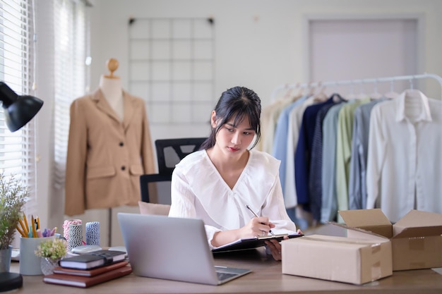 Mujeres comerciantes asiáticas jóvenes escribiendo órdenes de compra en línea de información del cliente en el portapapeles mientras trabajan para comprobar los datos en la computadora portátil y la entrega de envíos para el negocio de compras en línea en la oficina en casa