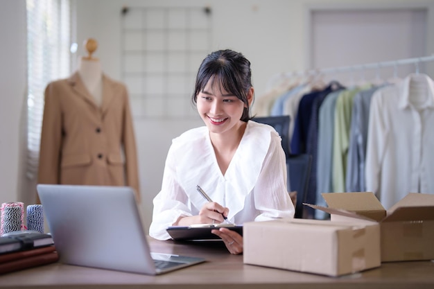 Mujeres comerciantes asiáticas jóvenes escribiendo órdenes de compra en línea de información del cliente en el portapapeles mientras trabajan para comprobar los datos en la computadora portátil y la entrega de envíos para el negocio de compras en línea en la oficina en casa