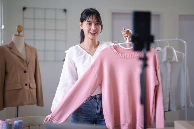 Foto mujeres comerciantes asiáticas jóvenes que usan su teléfono inteligente para grabar videos vendiendo ropa en línea para vlogger en las redes sociales mientras trabajan y envían entregas para negocios de compras en línea en la oficina en casa